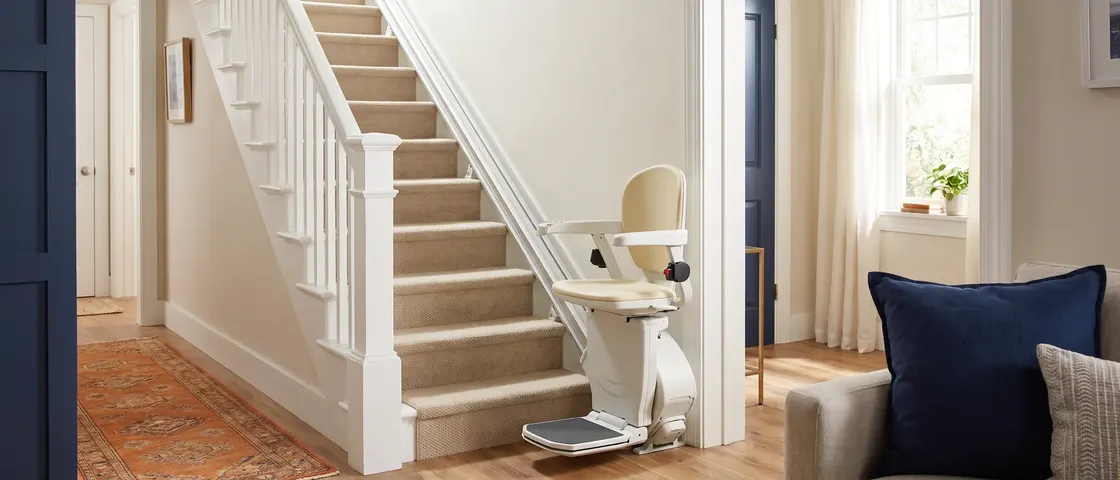 A white stair lift parked at the foot of a straight carpeted staircase