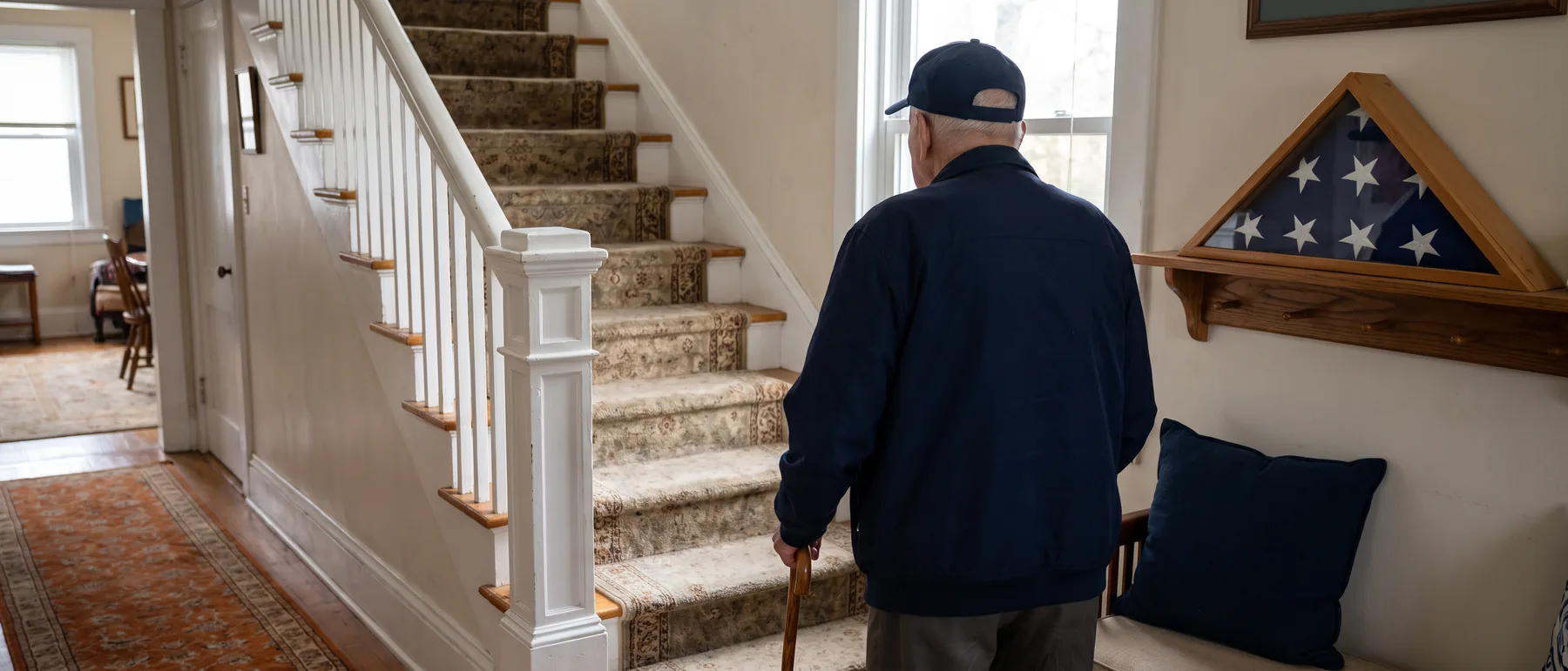 An older veteran with a cane at the foot of the stairs in his home, seen from behind